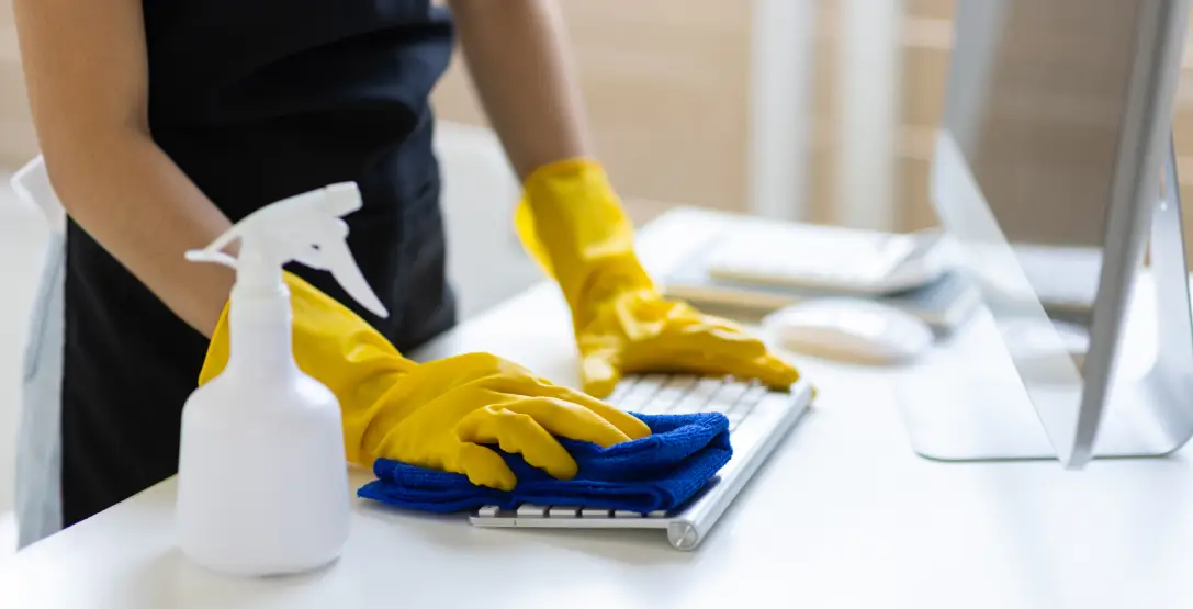A member of the Corporate Clean team, an office & janitorial cleaning services company, wipes down a computer keyboard sitting on a desk.
